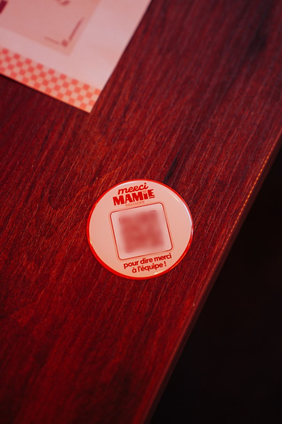 Une pastille Merci Mamie sur une table en bois rouge, avec un QR code flouté et la phrase « pour dire merci à l'équipe ! »